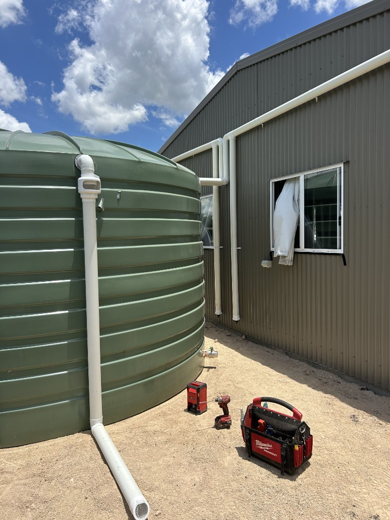 Green rainwater tank with PVC gutter connections beside a corrugated metal building